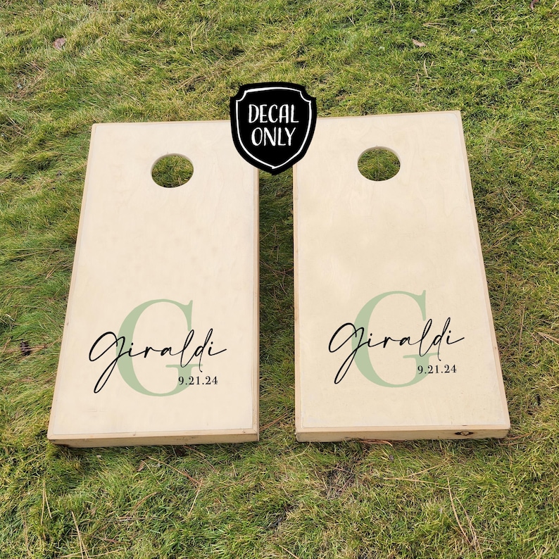 K&ouml;nnte beinhalten: Zwei h&ouml;lzerne Cornhole-Bretter mit einem gr&uuml;nen und schwarzen Aufkleber, auf dem "Giraldi 9.21.24" steht.