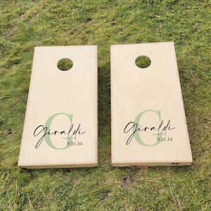 K&ouml;nnte beinhalten: Zwei h&ouml;lzerne Cornhole-Bretter mit einem gr&uuml;n-schwarzen Design. Die Bretter sind mit dem Namen "Giraldi" und dem Datum "9.21.24" personalisiert.