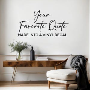 May include: A wall decal with the text "Your Favorite Quote MADE INTO A VINYL DECAL" in black script. The decal is displayed above a wooden console table with a vase, books, and a decorative chair.