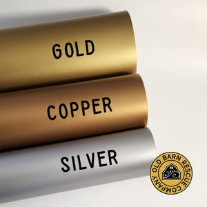Op de afbeelding: Drie rollen vinyl-kleeffolie in de kleuren goud, koper en zilver. De rollen zijn gelabeld met "GOLD", "COPPER" en "SILVER".