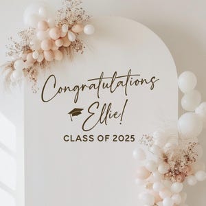 May include: A white arched backdrop with the words "Congratulations Ellie!" and "CLASS OF 2025" in dark brown script. The backdrop is decorated with white and blush-colored balloons and dried floral arrangements.