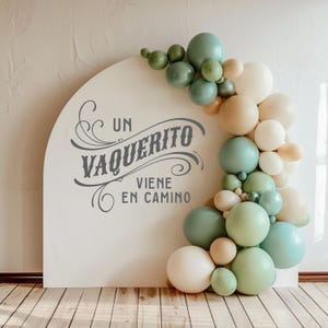 May include: A white arch with the text "Un Vaquerito Viene En Camino" in a decorative font. The arch is decorated with a garland of green, beige, and white balloons.