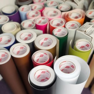 Puede incluir: Una colecci&oacute;n de rollos de vinilo en varios colores, incluyendo rosa, naranja, verde y blanco. Los rollos est&aacute;n bien enrollados, con los extremos mostrando el n&uacute;cleo interno y el logotipo de la marca. La imagen sugiere una tienda de manualidades o bricolaje.