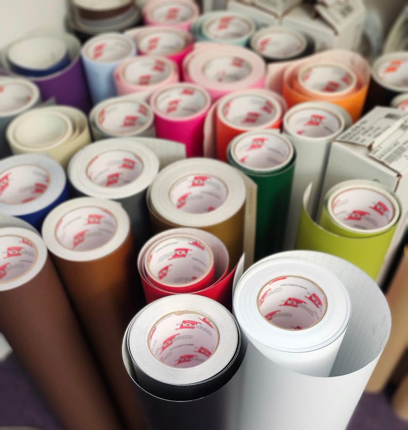 Puede incluir: Una colecci&oacute;n de rollos de vinilo en varios colores, incluyendo rosa, naranja, verde y blanco. Los rollos est&aacute;n bien enrollados, con los extremos mostrando el n&uacute;cleo interno y el logotipo de la marca. La imagen sugiere una tienda de manualidades o bricolaje.