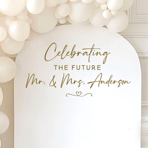 May include: A white arched sign with gold lettering that reads "Celebrating the Future Mr. & Mrs. Anderson." The sign is decorated with a heart and surrounded by white balloons, perfect for a wedding or engagement celebration.