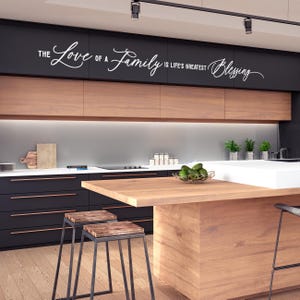 Op de afbeelding: Een moderne keuken met zwarte kasten en een houten aanrechtblad. De keuken heeft een witte muur met de tekst "The Love of a Family is Life's Greatest Blessing" geschreven in cursief.
