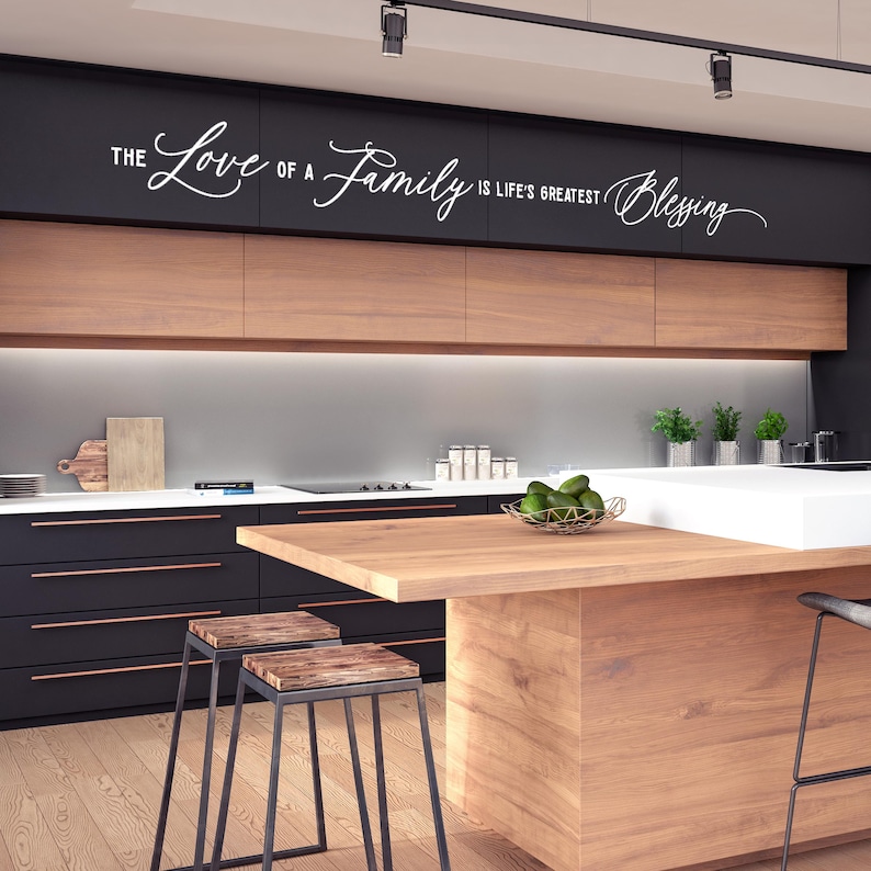 Op de afbeelding: Een moderne keuken met zwarte kasten en een houten aanrechtblad. De keuken heeft een witte muur met de tekst "The Love of a Family is Life's Greatest Blessing" geschreven in cursief.