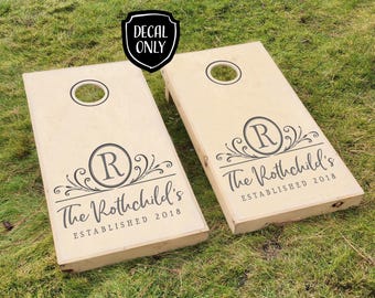 Könnte beinhalten: Zwei hölzerne Cornhole-Bretter mit individuellen Aufklebern. Die Aufkleber zeigen ein Monogramm mit dem Anfangsbuchstaben "R" mit einem floralen Design und dem Text "The Rothschild's Established 2018".