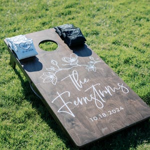 May include: A brown wooden cornhole game with white painted floral designs and the text "The Fernstroms 10.18.2024". Two white bean bags with the text "L&G 10.18.2024" and two black bean bags with the text "L&G 10.18.2024" are on the game.