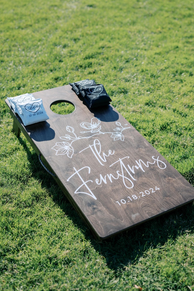 May include: A brown wooden cornhole game with white painted floral designs and the text "The Fernstroms 10.18.2024". Two white bean bags with the text "L&G 10.18.2024" and two black bean bags with the text "L&G 10.18.2024" are on the game.
