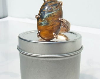 May include: A gold-coloured ring with a large, oval tiger's eye stone in shades of brown, gold, and blue. The ring has a textured band and four prongs holding the stone. The ring is displayed on a small, silver tin.