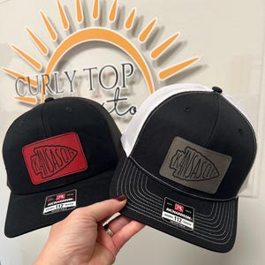 Könnte beinhalten: Zwei schwarze und weiße Trucker-Caps mit einem roten und grauen Pfeil-Patch, auf dem "Kansas City" steht. Die Caps haben ein weißes Netzrücken und eine schwarze Vorderseite.