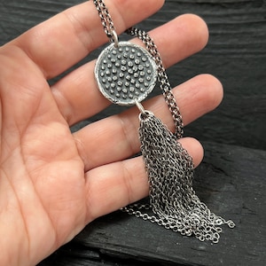 Peut inclure: Un collier argenté avec un pendentif circulaire texturé orné de petits points. Le pendentif est relié à une chaîne et à une longue pampille composée de plusieurs chaînes. Le bijou est présenté sur un fond sombre.