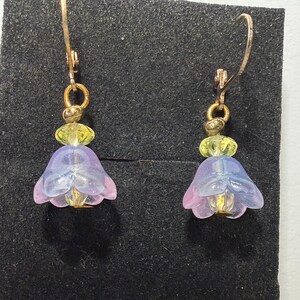 May include: A pair of drop earrings featuring gold-toned metal hooks and hardware. Each earring displays a small, translucent yellow bead positioned above a larger, bell-shaped, translucent purple and blue flower-shaped glass bead.