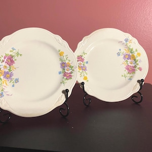 May include: Two off-white ceramic plates with scalloped edges and embossed detailing. Each plate features a floral pattern with pink, yellow, purple, and blue flowers arranged in clusters on the sides. The plates are held by black metal stands.