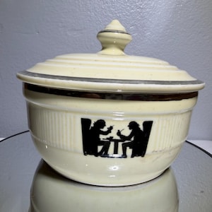 May include: A cream-colored ceramic lidded bowl featuring a dark brown horizontal band near the rim and a black silhouette graphic of two figures seated at a table. The lid has a central knob handle and a matching dark brown trim along the edge.