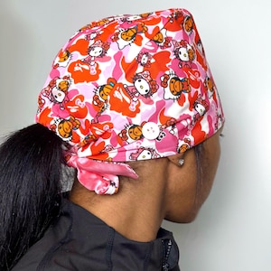 May include: A pink, red, and white patterned surgical scrub cap featuring a repeating design of a cartoon cat character. The cap has a button detail and a pink tie at the back. The cap is worn over a ponytail.