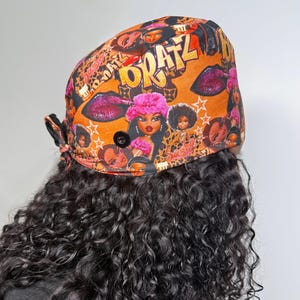 May include: A scrub cap featuring an orange background with a repeating pattern of Bratz doll characters, gold text reading Bratz, pink fuzzy hats, and white stars. The cap includes a black button on the side and a tie closure at the back.