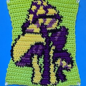 May include: A handmade crochet piece featuring a stylized mushroom design. The mushroom is rendered in shades of yellow, purple, and black, set against a bright green background. The edges of the piece are finished with a neat, even stitch.