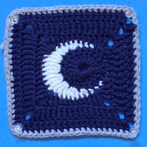 Peut inclure: Un carré crocheté avec un motif de croissant de lune blanc sur un fond bleu foncé. Le carré a une bordure grise et est fixé à une surface bleue avec des punaises en plastique transparent.