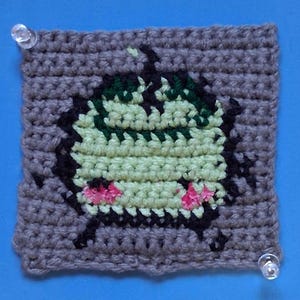 May include: A crocheted square with a green, black, and pink design of a cartoon character. The character has a green body, black eyes, and pink cheeks.