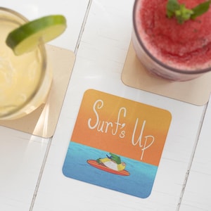 May include: A square coaster with a yellow, orange, and blue sunset design. The text "Surf's Up" is written in white cursive. A cartoon parrot is riding a surfboard in the ocean.