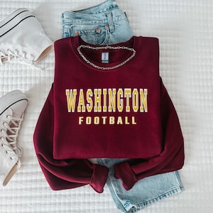 May include: A burgundy sweatshirt featuring the text "WASHINGTON FOOTBALL" in yellow lettering. The image includes blue denim jeans and white trainers, offering a relaxed, athletic style.