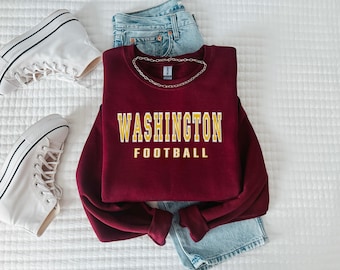 Könnte beinhalten: Ein weinrotes Sweatshirt mit dem gelben Schriftzug "WASHINGTON FOOTBALL". Dazu eine blaue Jeans und weiße Turnschuhe, die einen lässigen, sportlichen Look erzeugen.