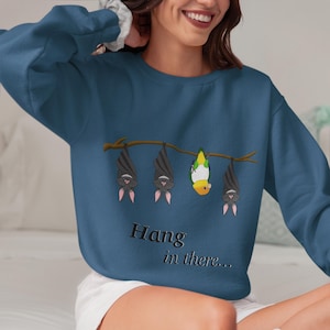 Op de afbeelding: Een blauw sweatshirt met een afbeelding van vleermuizen die ondersteboven aan een tak hangen, met een gele en groene papegaai die ernaast ondersteboven hangt. De tekst "Hang in there..." staat onder de afbeelding.