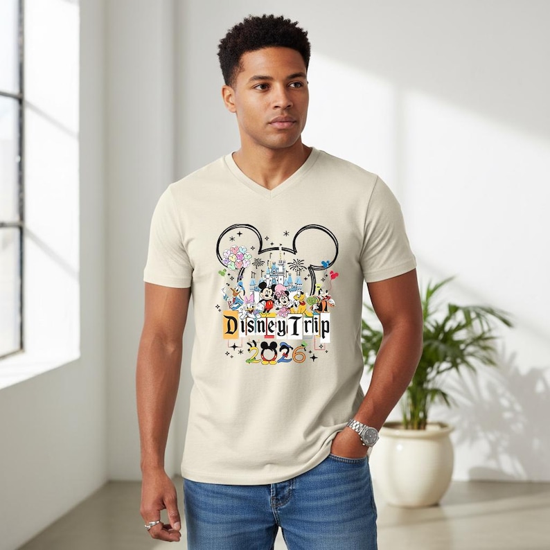 A man wearing a white t-shirt with a Disney-themed graphic and text, standing in a room with a potted plant in the background.