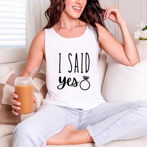A woman sitting on a couch, wearing a white tank top with the text "I said yes" and holding a drink.