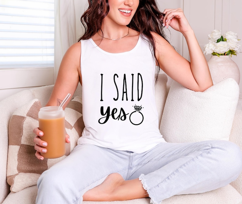 A woman sitting on a couch, wearing a white tank top with the text "I said yes" and holding a drink.