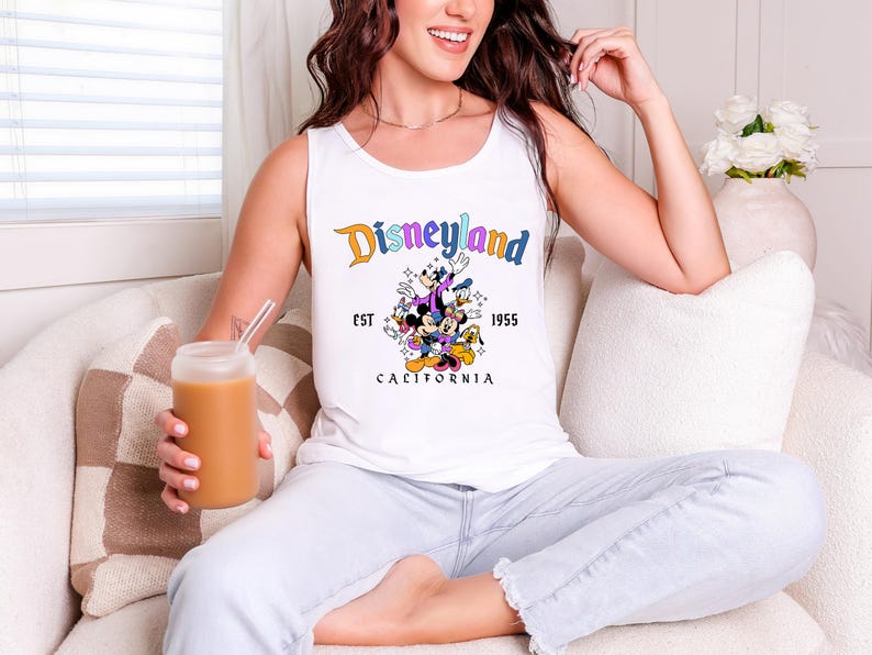 A woman sitting on a couch, wearing a white tank top with a Disney logo, and holding a drink.