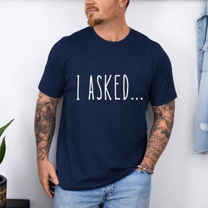 A man wearing a navy blue t-shirt with the text "I asked..." standing in front of a white wall.
