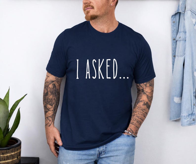 A man wearing a navy blue t-shirt with the text "I asked..." standing in front of a white wall.