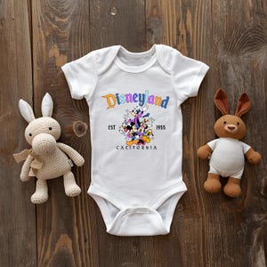 A white onesie with Disneyland text and a rabbit toy is placed on a wooden surface, with a stuffed rabbit toy to the left and a stuffed animal to the right.