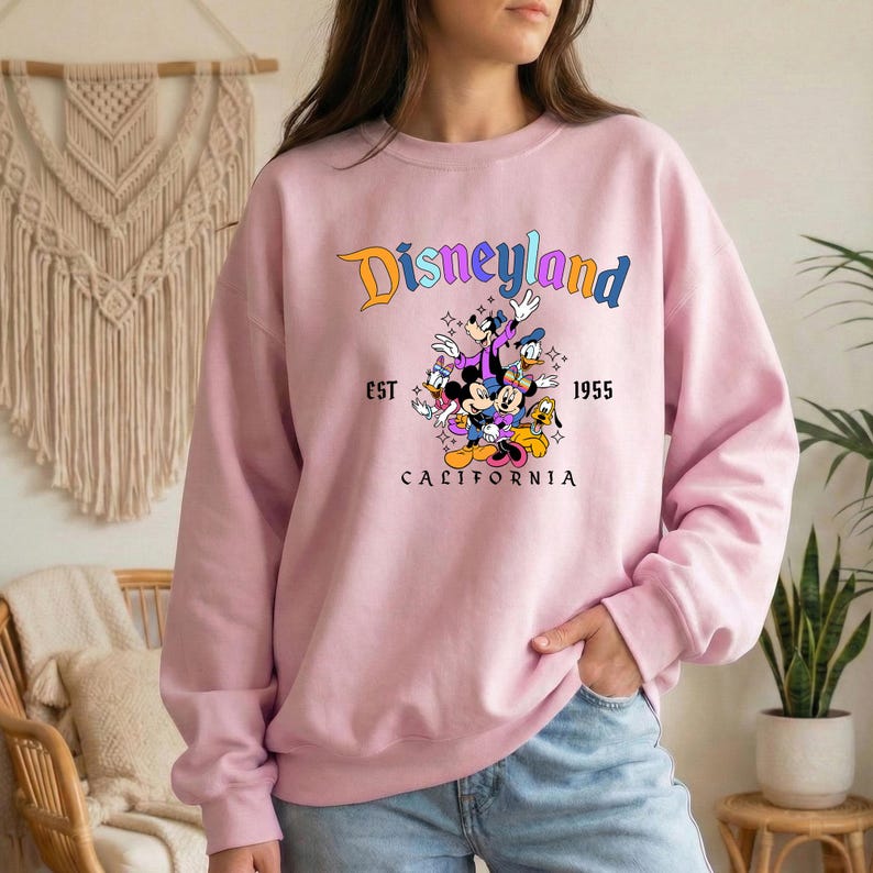 A woman wearing a pink sweatshirt with a Disneyland logo and text, standing in front of a wall with a macrame decoration.