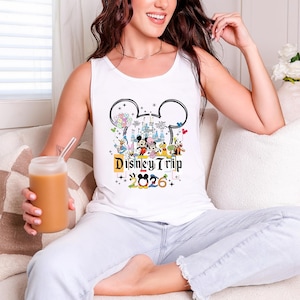 A woman sitting on a couch, wearing a Disney Trip tank top and holding a drink, smiling and looking at the camera.