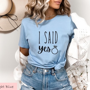 A woman wearing a light blue t-shirt with the text "I said yes" printed on it, paired with denim shorts and a white cardigan.