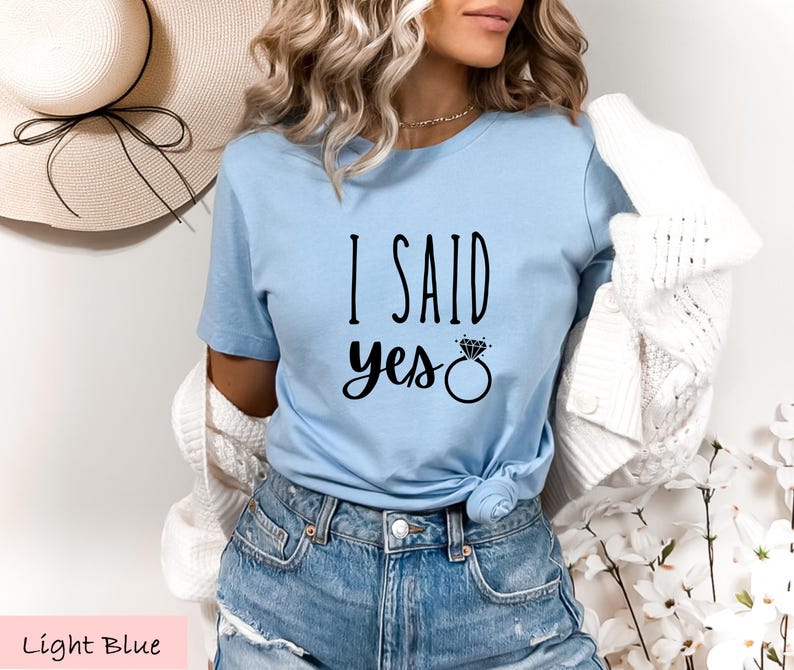 A woman wearing a light blue t-shirt with the text "I said yes" printed on it, paired with denim shorts and a white cardigan.