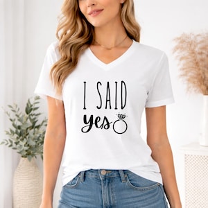 A woman wearing a white t-shirt with the text "I said yes" printed on it, standing in front of a window with a plant and a vase.