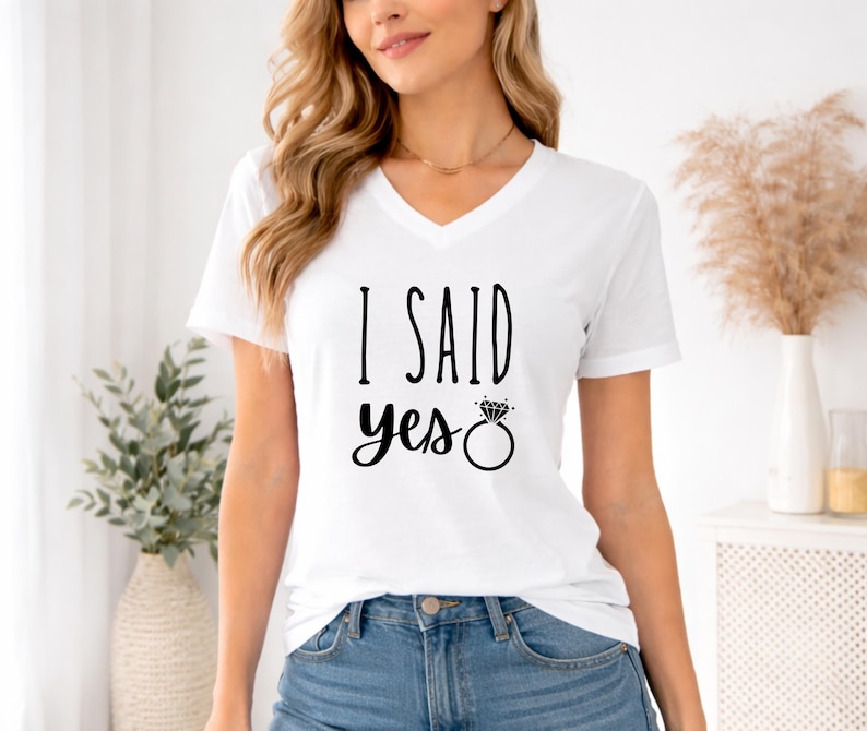 A woman wearing a white t-shirt with the text "I said yes" printed on it, standing in front of a window with a plant and a vase.