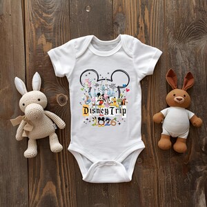 A white baby onesie with Disney Trip graphics and the text "Disney Trip" is placed between a stuffed rabbit and a stuffed bear on a wooden surface.
