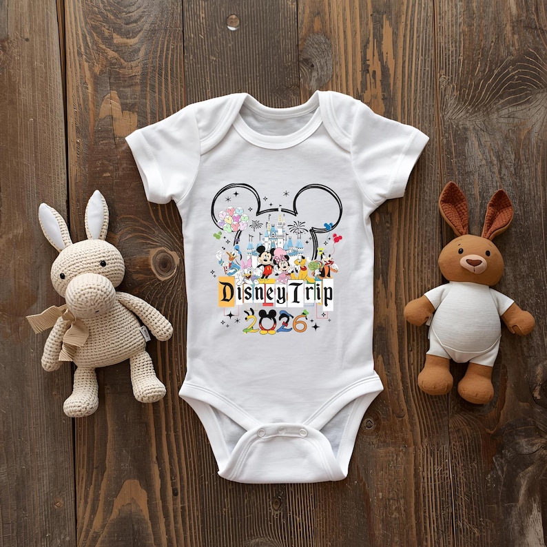 A white baby onesie with Disney Trip graphics and the text "Disney Trip" is placed between a stuffed rabbit and a stuffed bear on a wooden surface.