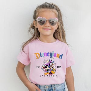A young girl wearing a pink t-shirt with a Disney theme graphic and heart-shaped sunglasses, smiling at the camera.