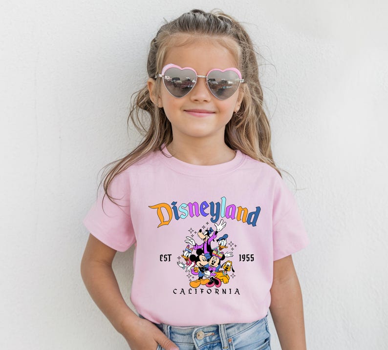 A young girl wearing a pink t-shirt with a Disney theme graphic and heart-shaped sunglasses, smiling at the camera.
