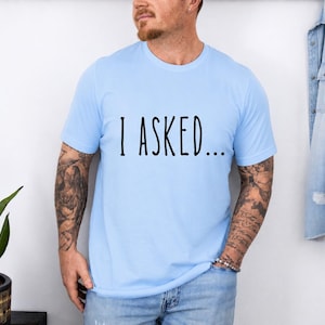 A man wearing a light blue t-shirt with the text "I asked..." standing in front of a white wall.