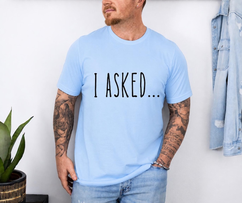 A man wearing a light blue t-shirt with the text "I asked..." standing in front of a white wall.