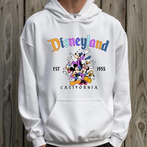 A person wearing a white hoodie with a graphic of Disney characters and text that says "Disneyland California".