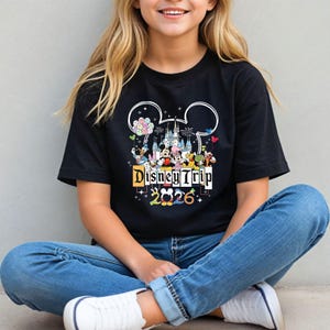 A smiling young woman wearing a black t-shirt with a Disney-themed graphic and text, sitting on the ground.